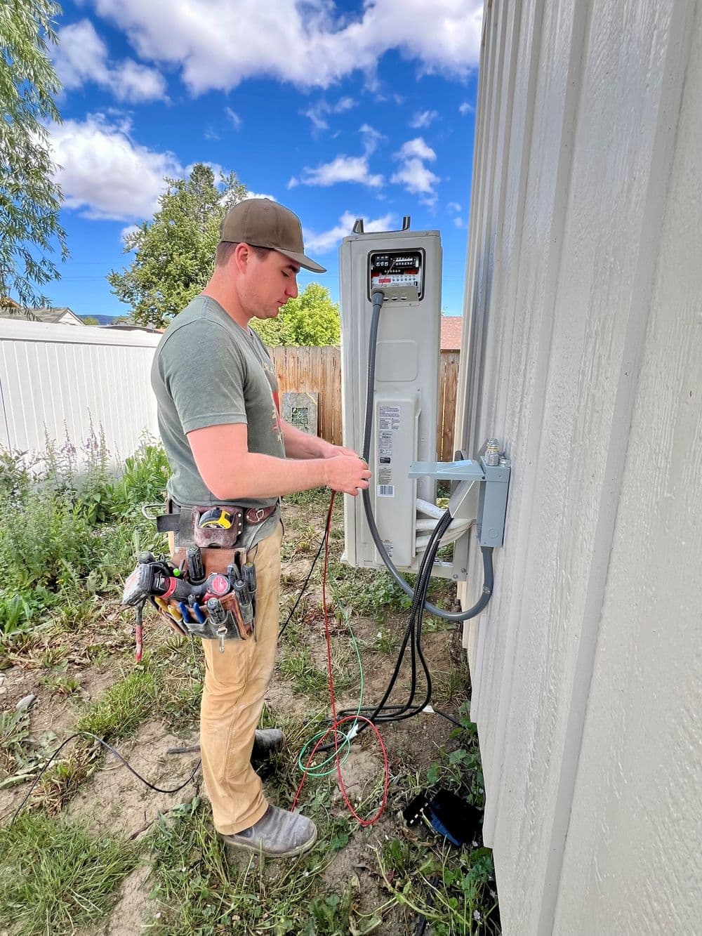 Electrician working on an outdoor electrical panel, wearing tools, in a residential yard.